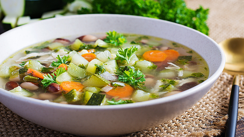 Das Bild zeigt einen Teller voller Gemüsesuppe mit Hühnchenfilet auf einem rustikalen Holztisch. 