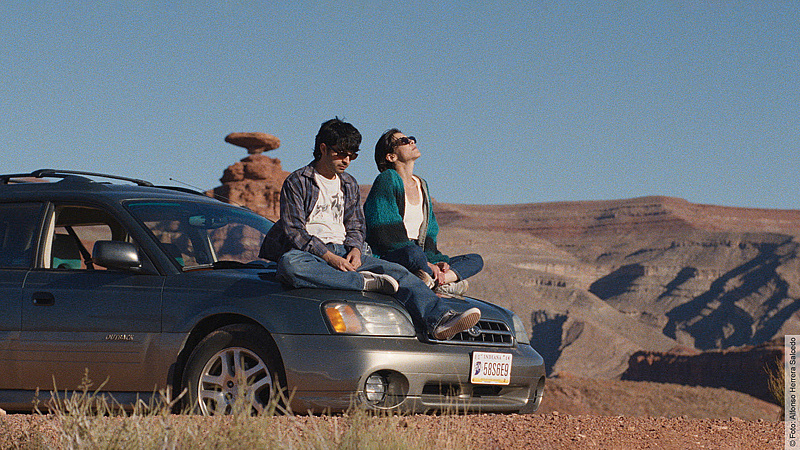 Ein Mann und eine Frau sitzen nebeneinander auf der Motorhaube eines dunkelgrauen Kombis (Subaru Outback) inmitten einer weiten Wüstenlandschaft. Der Mann trägt ein kariertes Hemd, Jeans sowie eine Sonnenbrille und blickt nach unten. Die Frau neben ihm trägt eine blau-schwarz gestreifte Strickjacke, hat ebenfalls eine Sonnenbrille auf und neigt den Kopf nach hinten in die Sonne. Im Hintergrund erstreckt sich eine rötliche Canyon- und Felslandschaft mit einer markanten pilzförmigen Felsformation unter einem klaren, blauen Himmel.